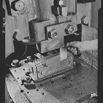 Conversion. Toy factory. Between the heavy punch and the die, a vital war product is being turned out by a press that once produced onlymetal toys and movie projectors. U-shaped object is part of radio filter box essential to the army. Keystone